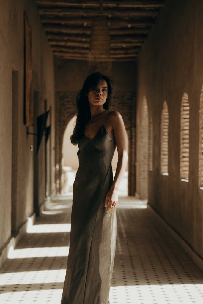 A stylish woman in a long dress stands in a sunlit hallway with architectural shadows cast.