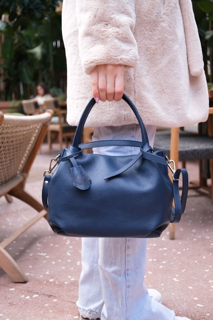 A woman in a white jacket stands holding a blue leather handbag, showcasing elegant fashion style.