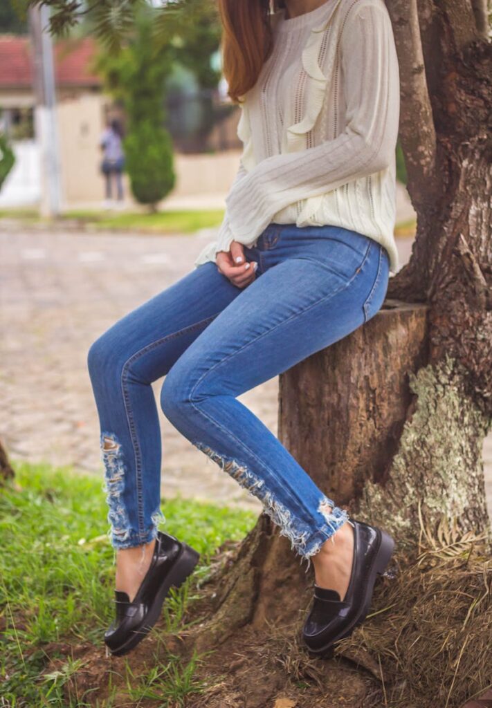 Fashionable woman sitting on a tree trunk, showcasing urban style with ripped jeans and a cozy sweater.