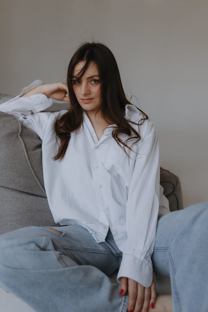 A woman in a white shirt casually poses on a sofa, exuding elegance and style.