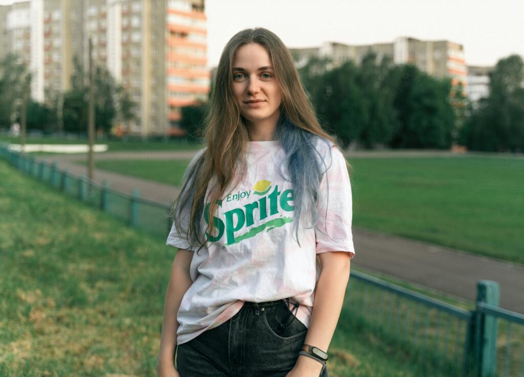 Young woman with long brown hair in a park, wearing a Sprite t-shirt and black jeans.