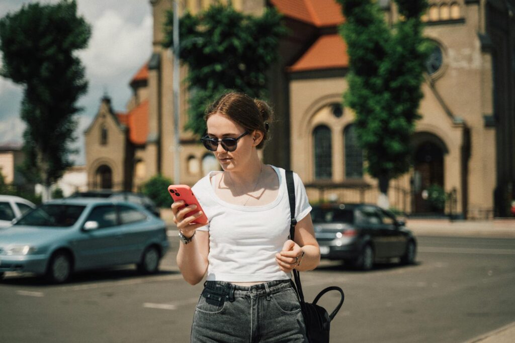 Young woman with sunglasses using smartphone in sunny urban street with historic architecture.