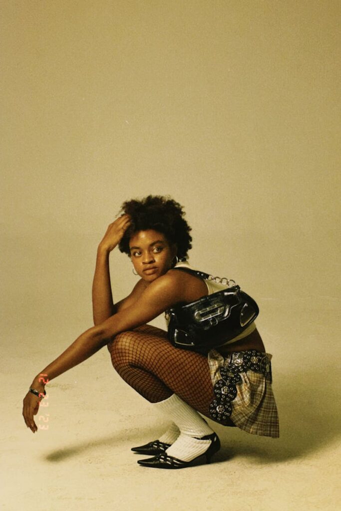 Black model with short hair squatting in a stylish outfit with a yellow background.