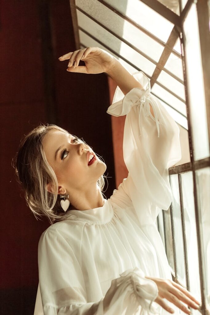 A stylish woman in a white blouse poses thoughtfully by a sunlit window indoors.