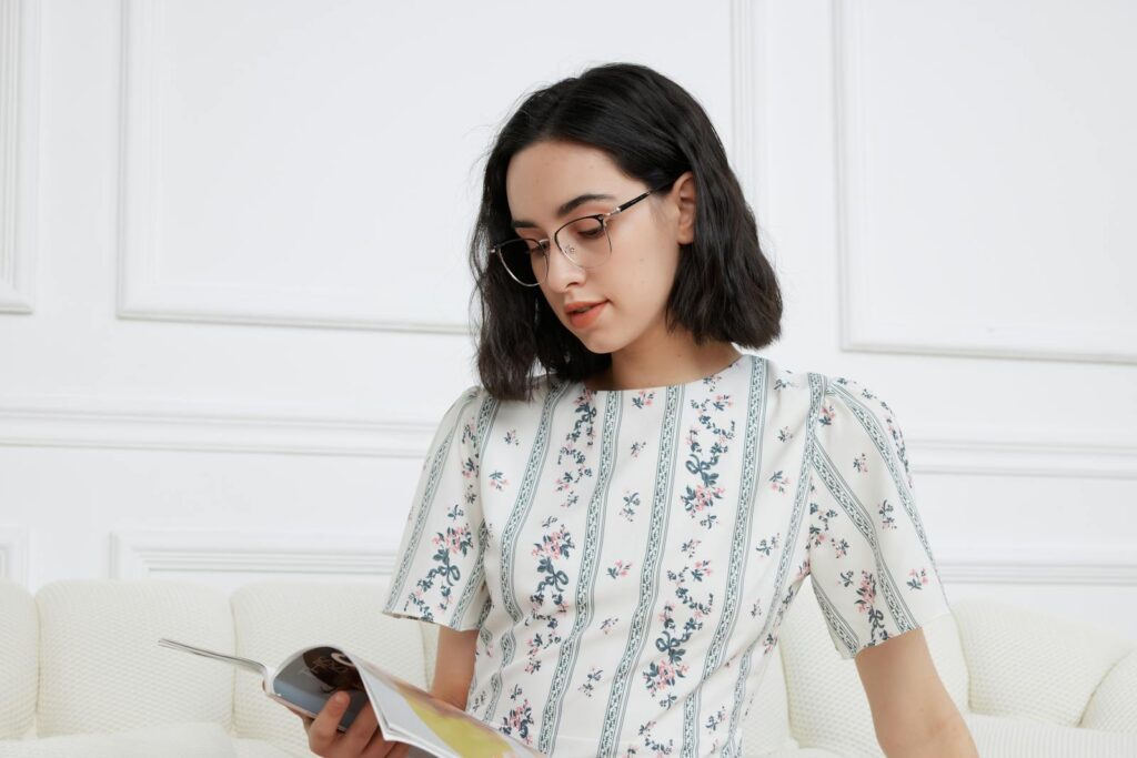 Portrait of a woman in glasses reading a magazine indoors wearing a floral dress.