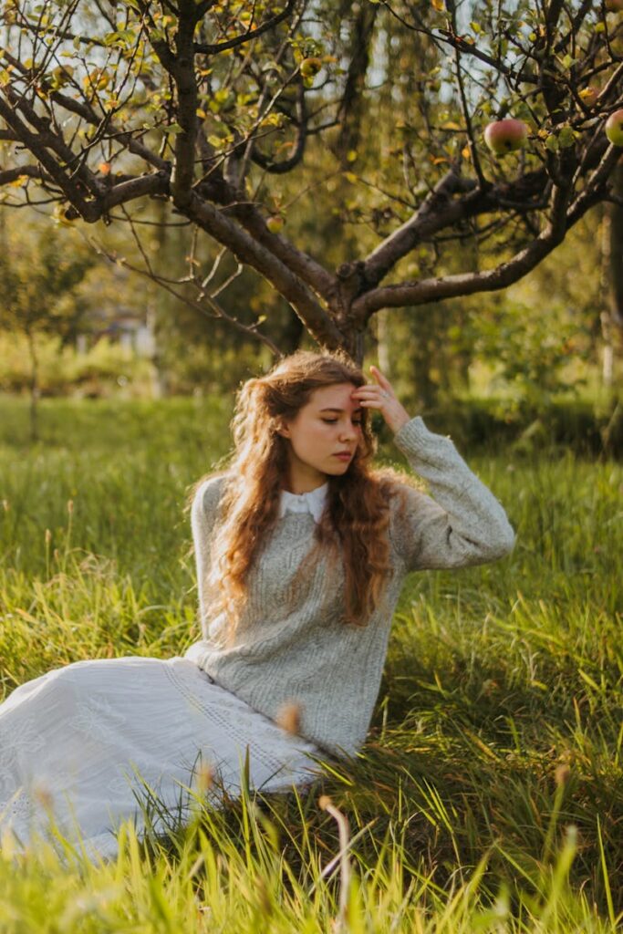 Young woman in cozy sweater sitting under an apple tree in a lush green orchard, enjoying a sunny day.