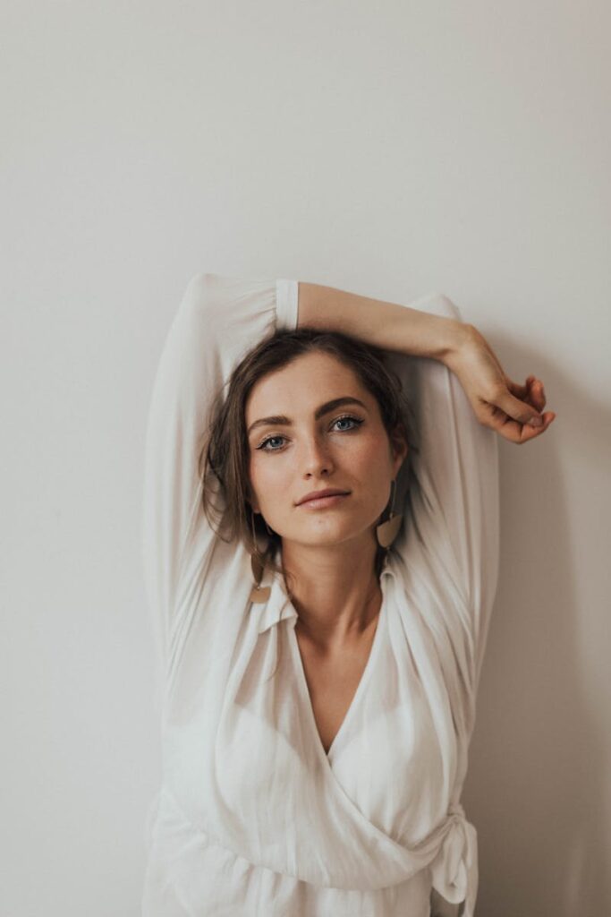 A beautiful woman in a white shirt poses casually against a white wall, exuding elegance and calm.