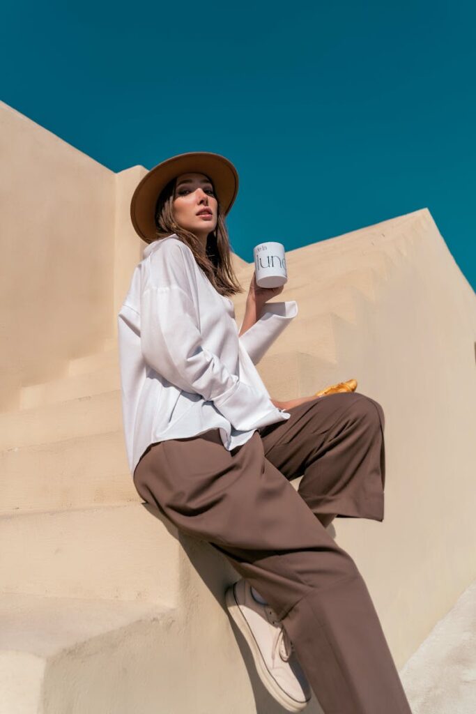 Fashionable woman in wide-brim hat holding coffee cup, enjoying sunny day on steps.