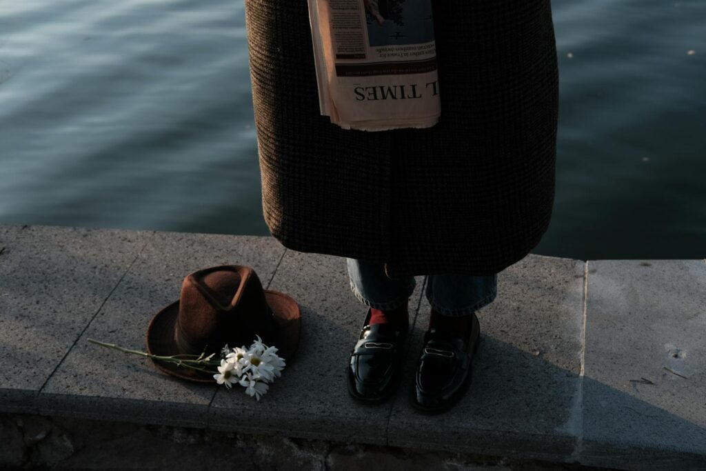 A person stands by the water with a hat, flowers, and a newspaper, evoking a cinematic mood.