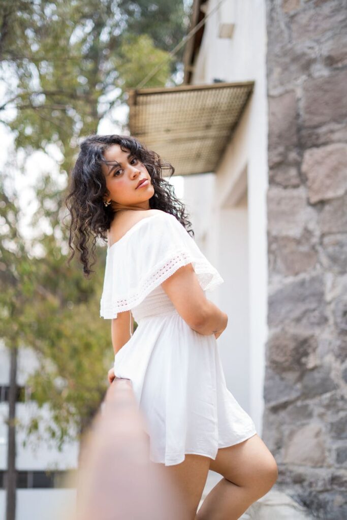 Elegant young woman with curly hair wearing a white dress, posing confidently against outdoor backdrop.