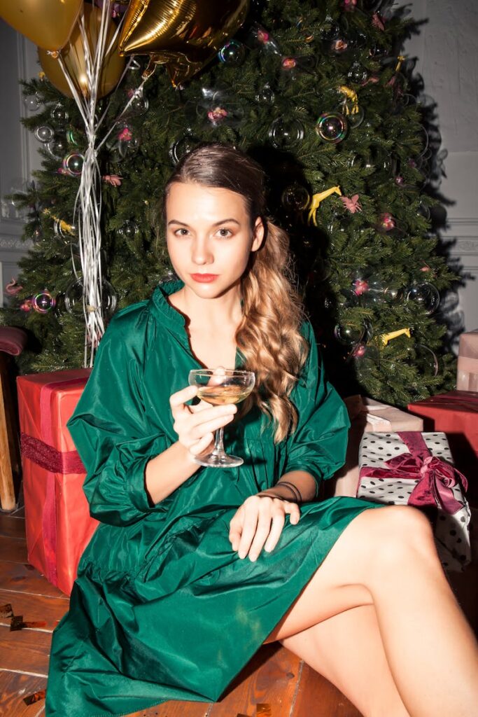 Woman in green dress holding champagne glass by Christmas tree at a festive indoor celebration.