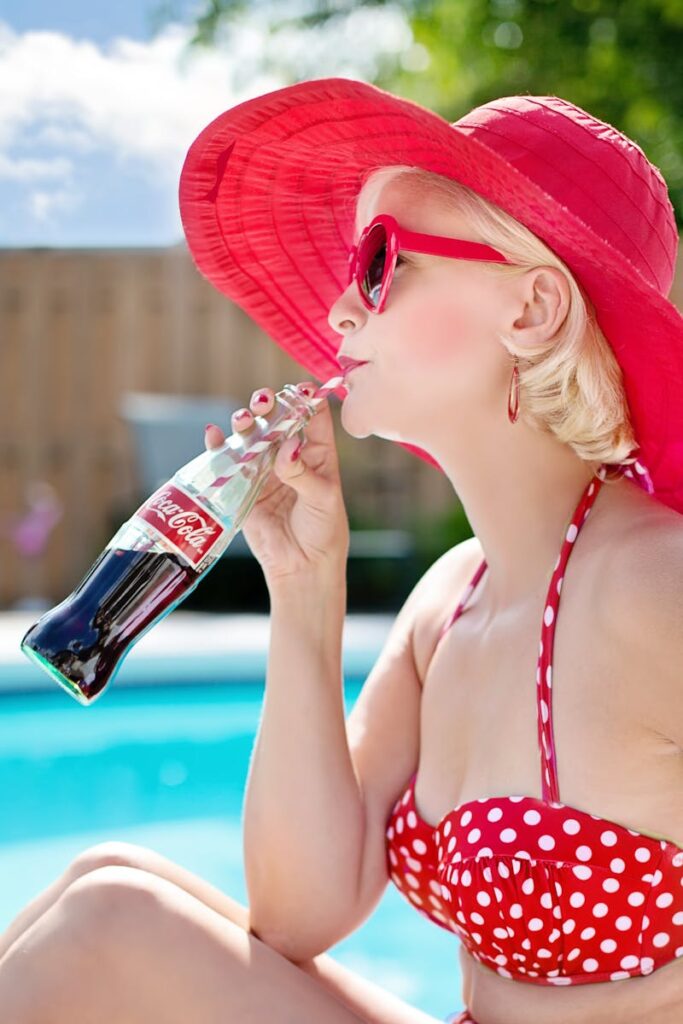 Chic woman sipping Coca-Cola by the pool in a red hat and polka dot bikini.