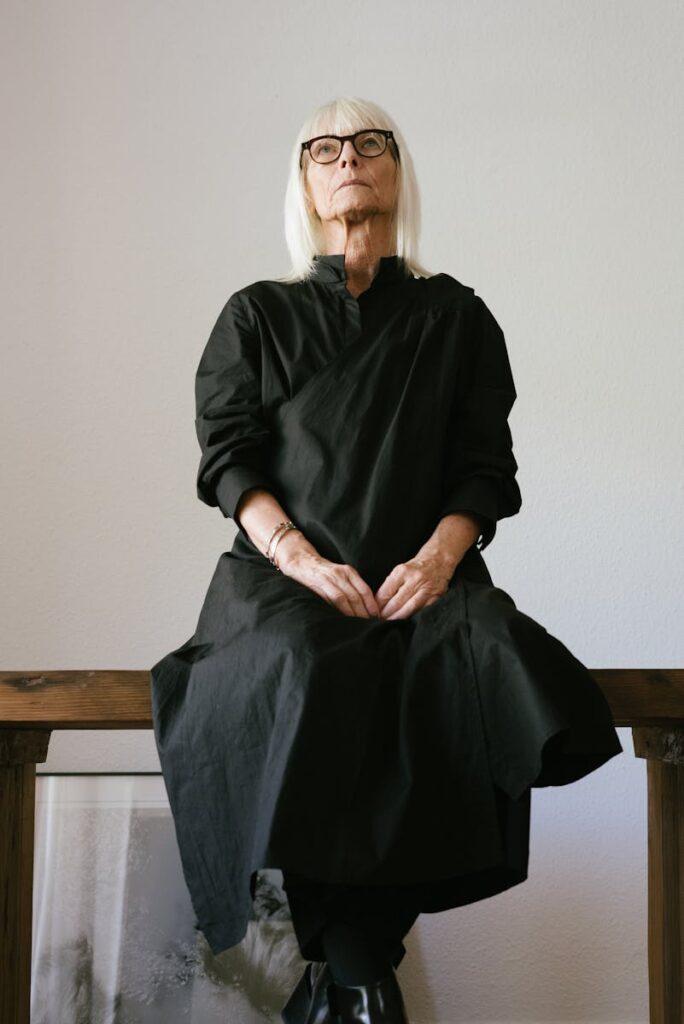 Senior woman with white hair sits indoors in a chic black gown, exuding elegance.