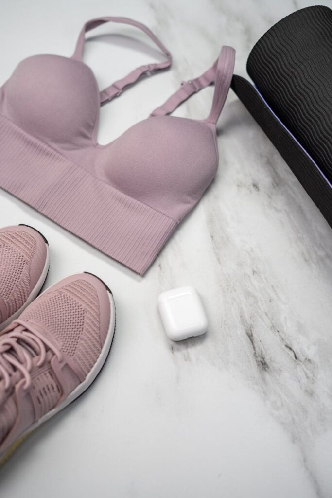 Pastel pink sports bra, sneakers, and yoga mat with earphones on marble.