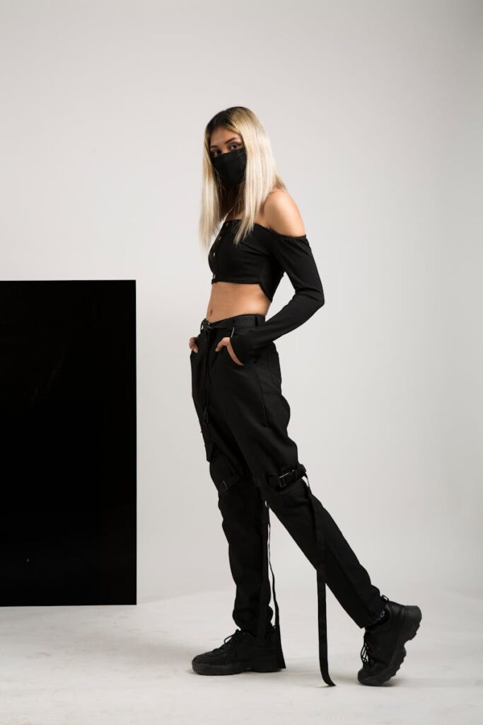 Fashionable young woman in black outfit and mask posing in studio setting.