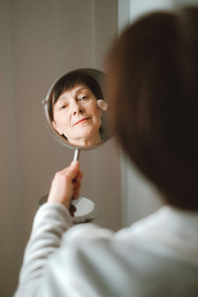 A senior caucasian woman smiling and applying makeup in front of a handheld mirror indoors.