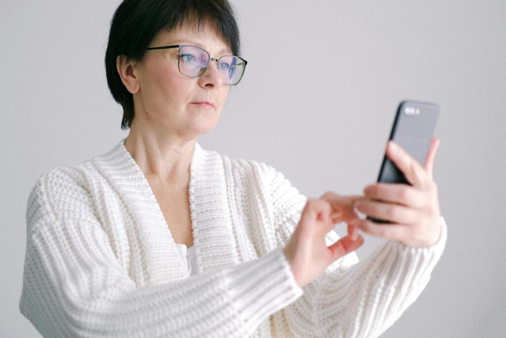 Elderly woman in glasses using a smartphone indoors, wearing a white sweater.