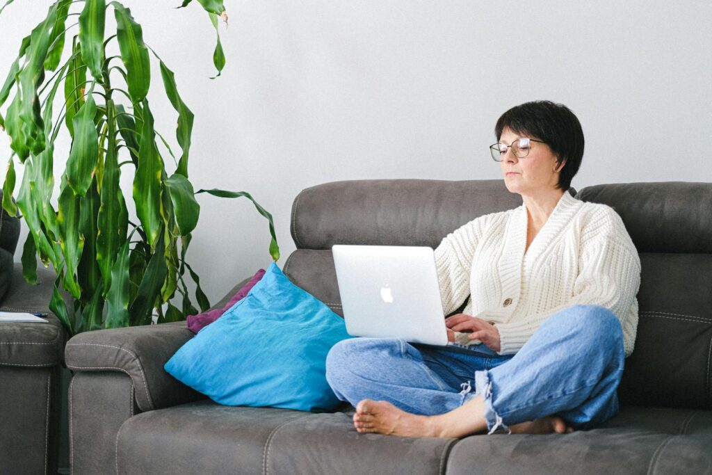 Elderly woman in a cardigan using a laptop while sitting on a couch surrounded by home decor.