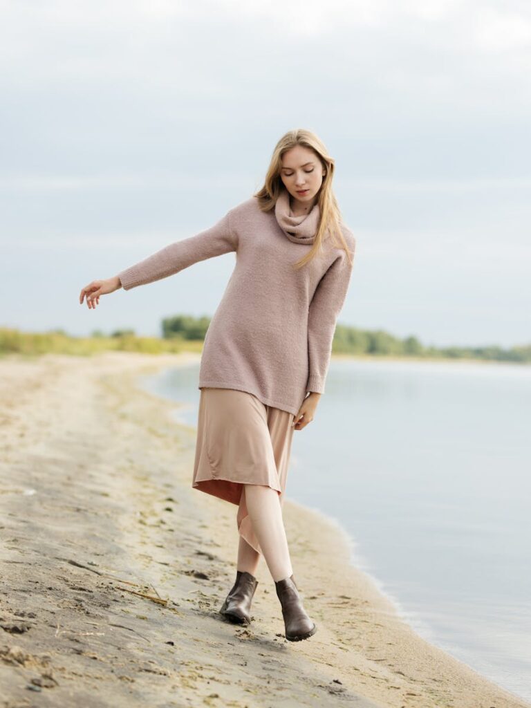 Woman in a sweater and skirt walking gracefully along a serene lakeside beach.
