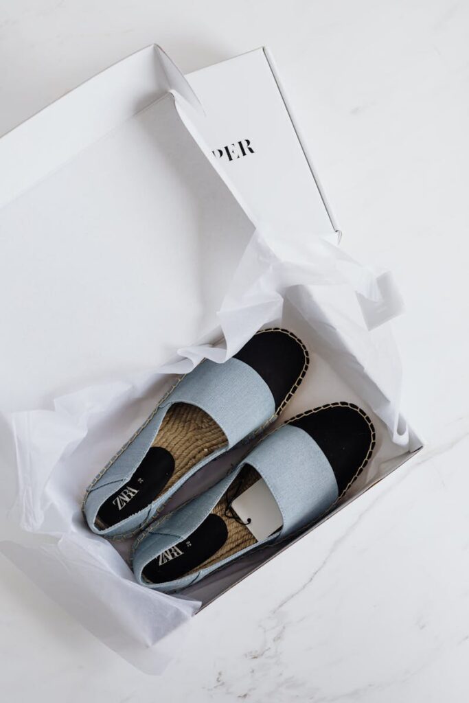 Top view of fashionable espadrilles in an open white cardboard box on a marble surface.