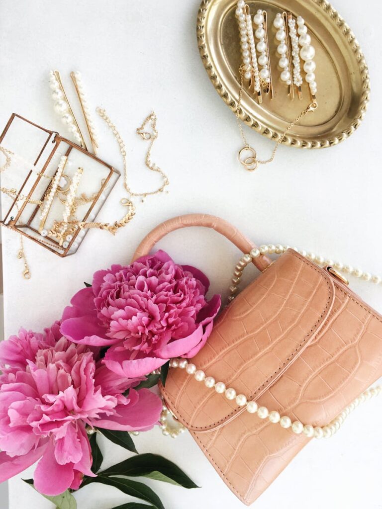 Chic flat lay design featuring accessories, jewelry, pink peonies, and a stylish handbag on a white surface.