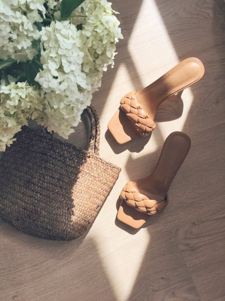Stylish summer sandals and a woven bag next to white hydrangeas in sunlight on wooden floor.