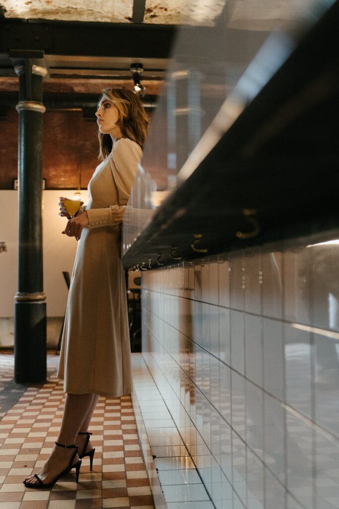 Elegant woman in a stylish dress holding a drink, standing indoors in a bar with a sophisticated ambiance.
