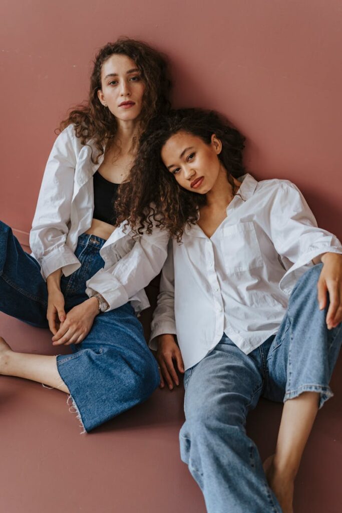 Two women with curly hair wearing denim and white shirts posed stylishly against a plain background.