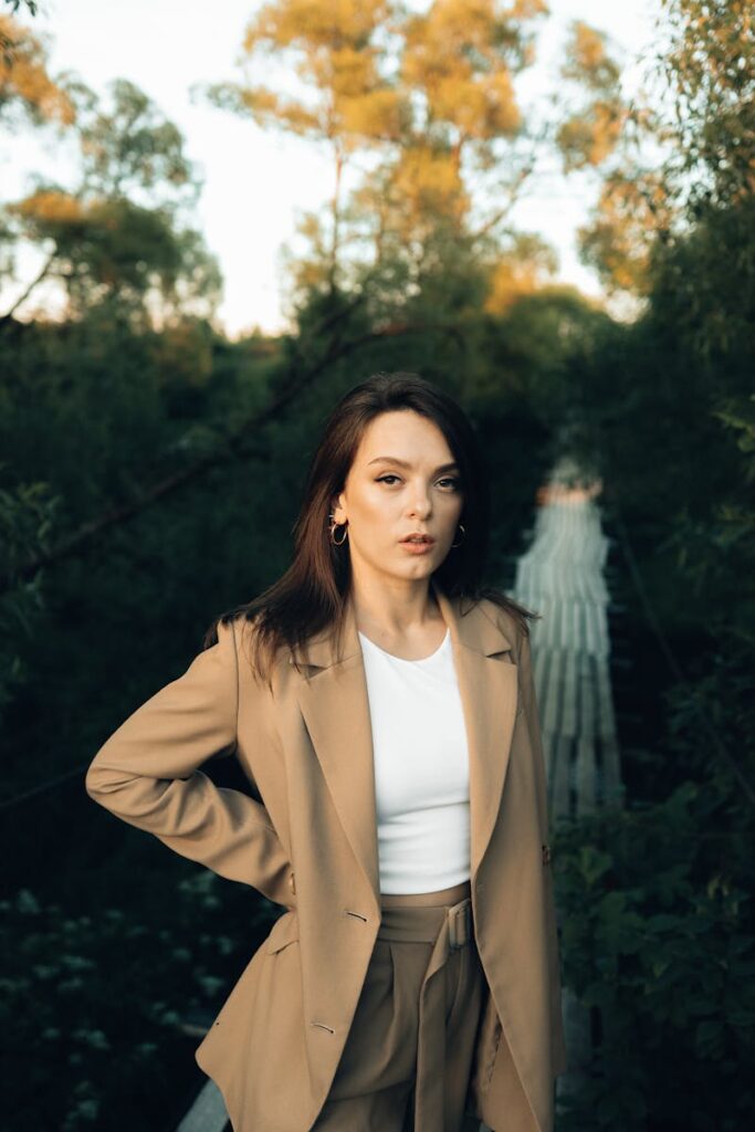 Fashionable woman in tan blazer stands confidently on a bridge surrounded by nature.