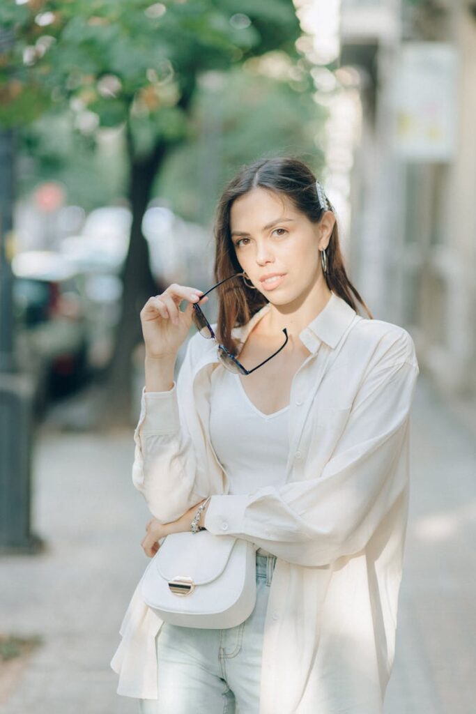 Fashionable woman in white blouse holding sunglasses outdoors on a sunny day.