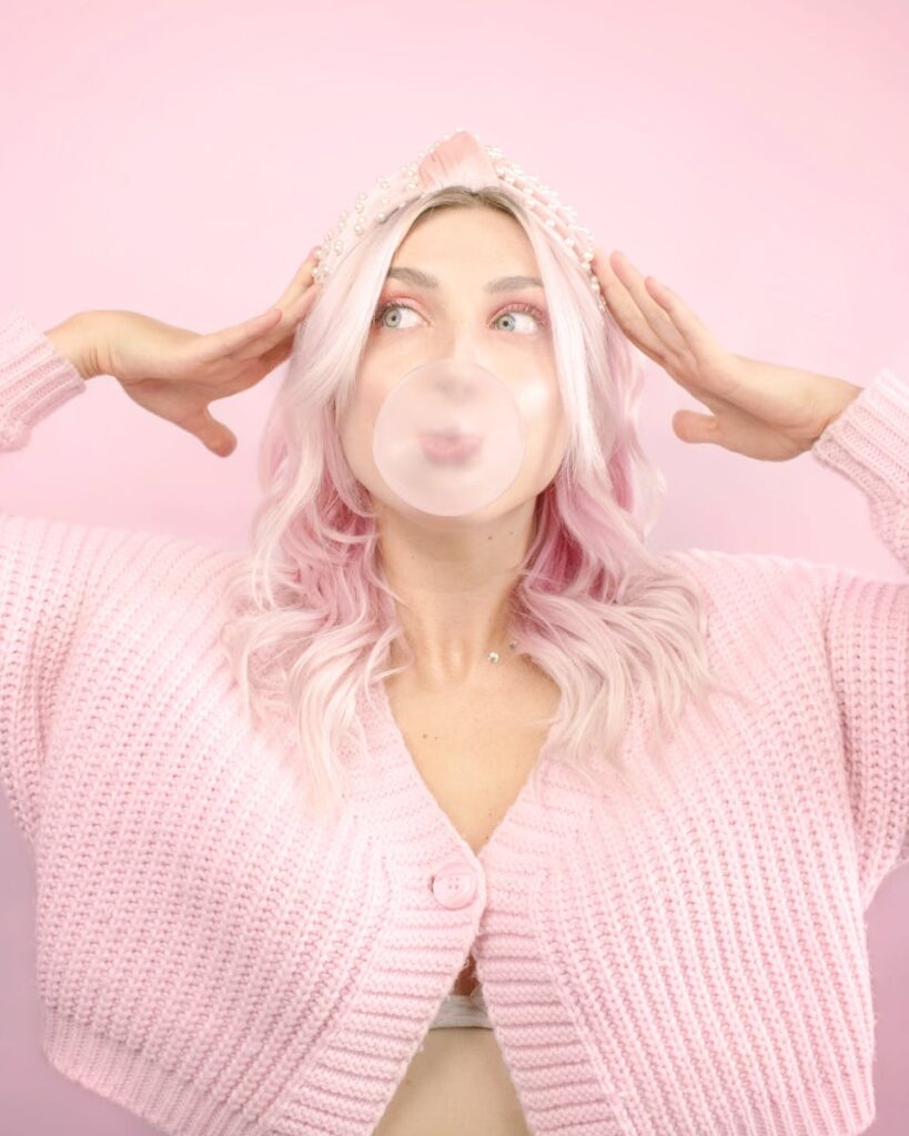 A woman with pink hair and cardigan playfully poses with bubble gum on a pastel pink background.