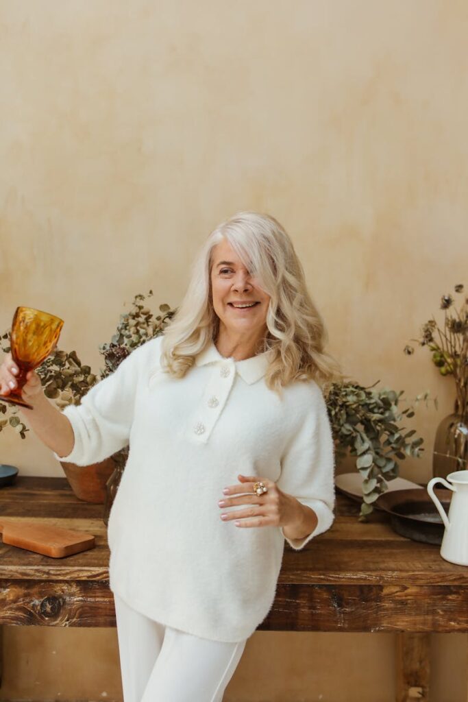 Smiling elderly woman holding a glass indoors, surrounded by dried flowers.