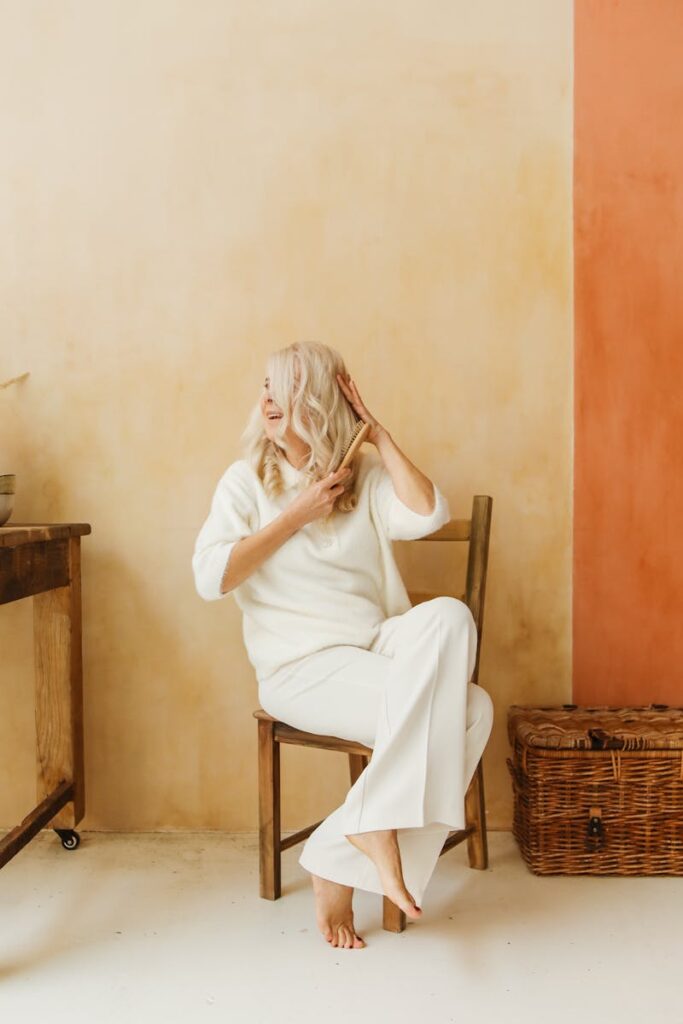 Graceful senior woman with long hair sitting barefoot on a wooden chair in a minimalist setting.