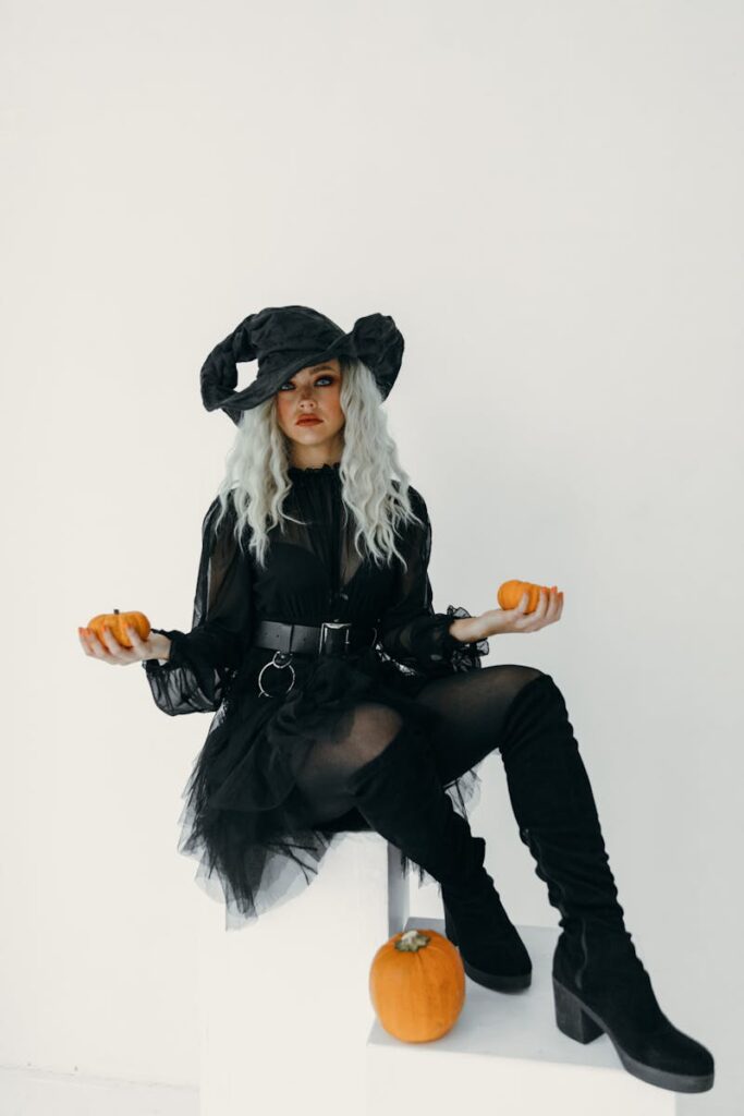 A fashionable woman in a witch costume poses with pumpkins indoors, perfect for Halloween themes.