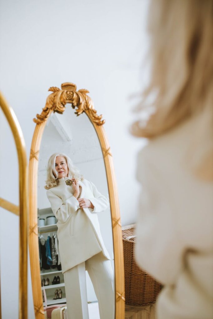 Senior woman in white outfit looking at her reflection in a vintage mirror inside a stylish room.