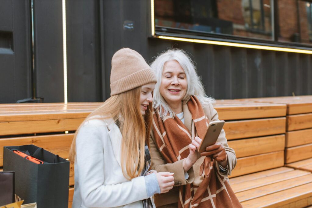 A senior woman sharing a moment with a teenager while looking at a smartphone outdoors on a wooden bench.