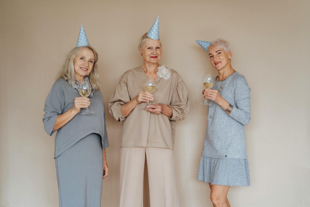 Three joyful senior women in party hats with glasses of wine celebrating indoors.