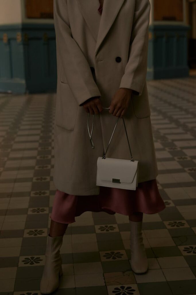 Elegant woman in a beige coat holding a clutch in an indoor tiled location.