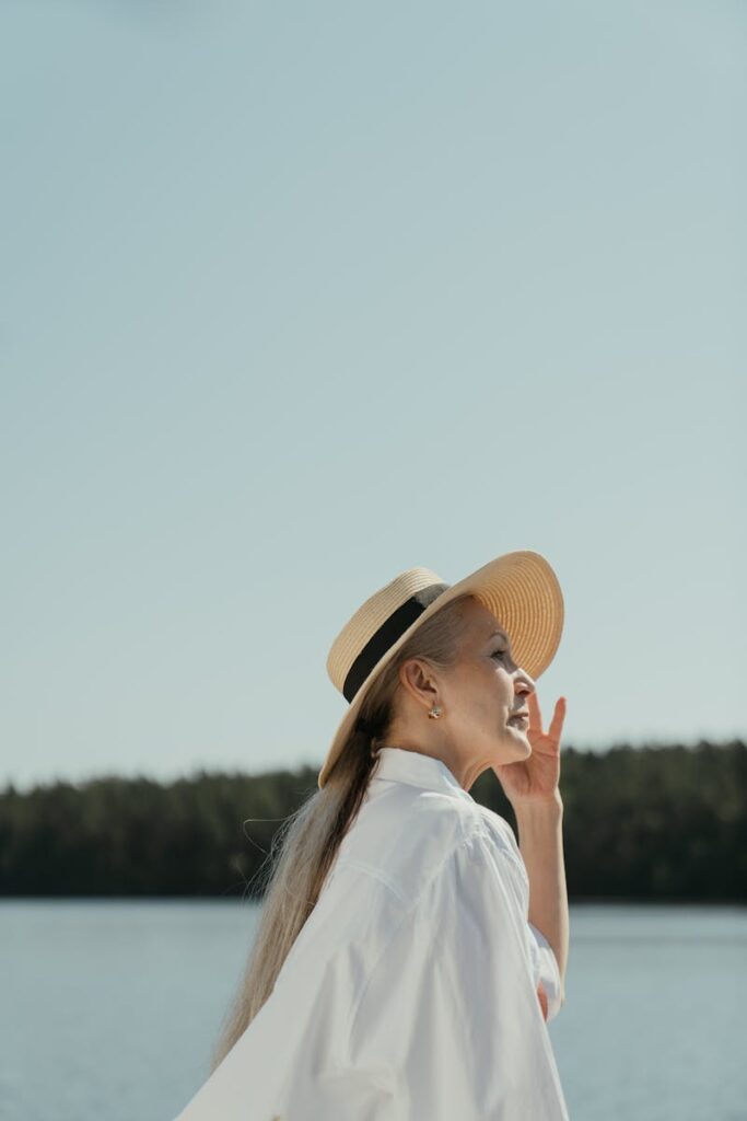 Side profile of a stylish elderly woman in a sun hat, enjoying the serene view by a lake under a clear sky.