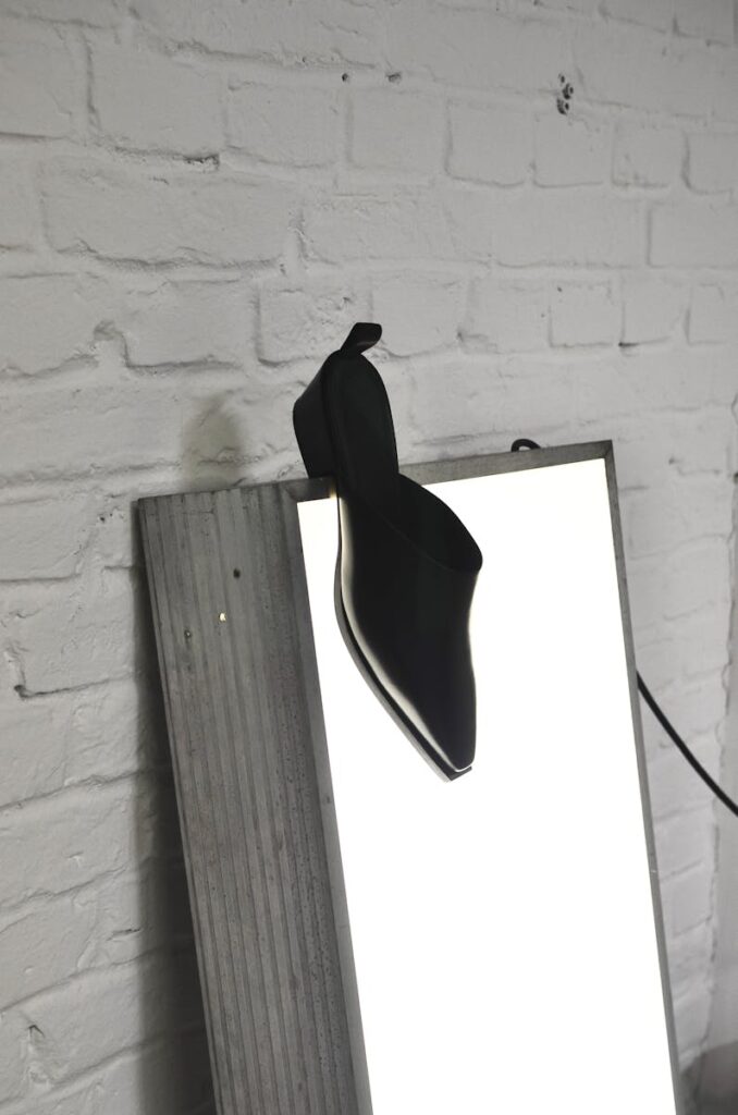 Elegant black mule shoe displayed on a brightly lit panel against white brick wall.