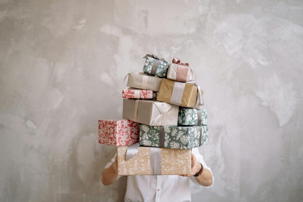 Person hiding behind a tall stack of gift-wrapped boxes against a gray wall, indoors.