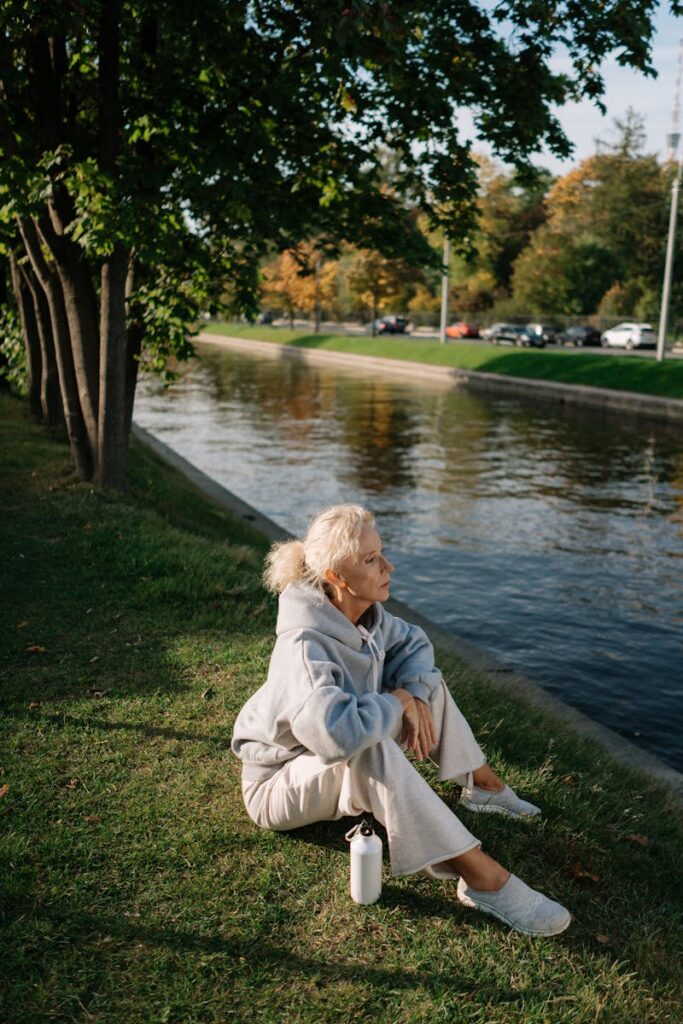 Elderly woman in casual wear sitting by a tranquil river outdoors with autumn foliage.