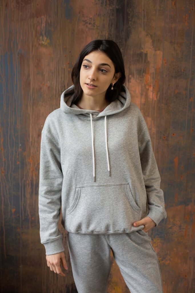 Emotionless young ethnic female wearing gray hoodie and pants standing with hand in pocket against weathered wooden wall and looking away
