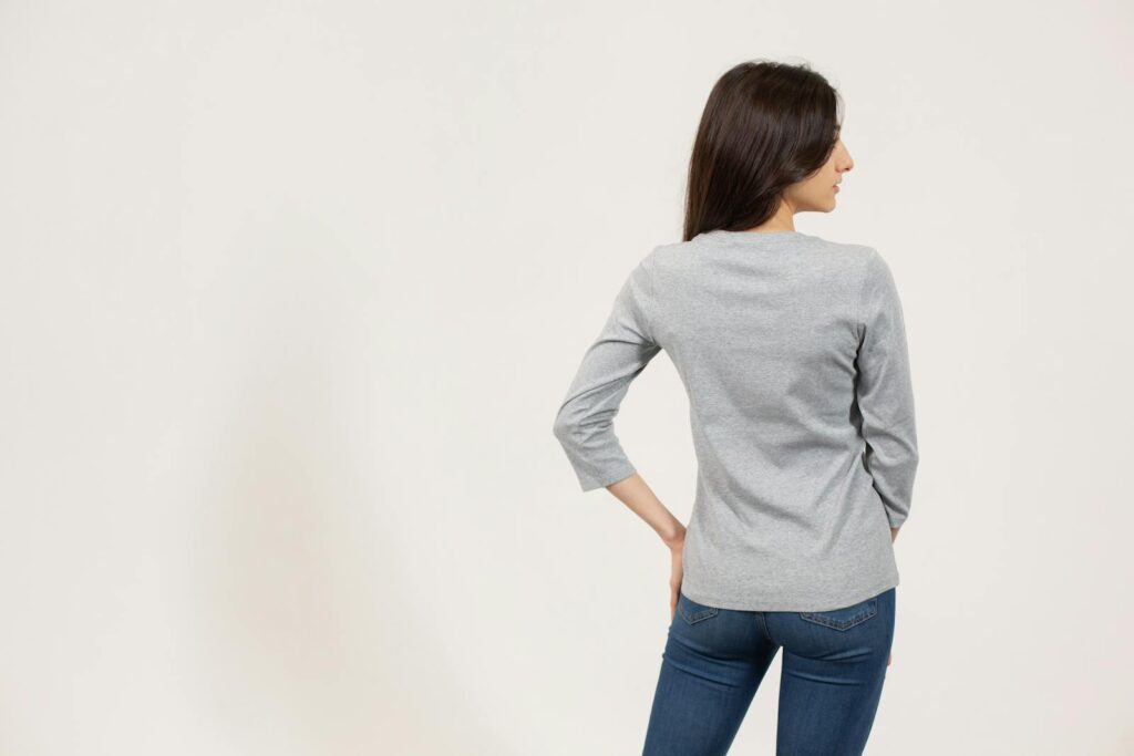 Back view of female model in jeans and gray long sleeve t shirt standing against white background and looking away