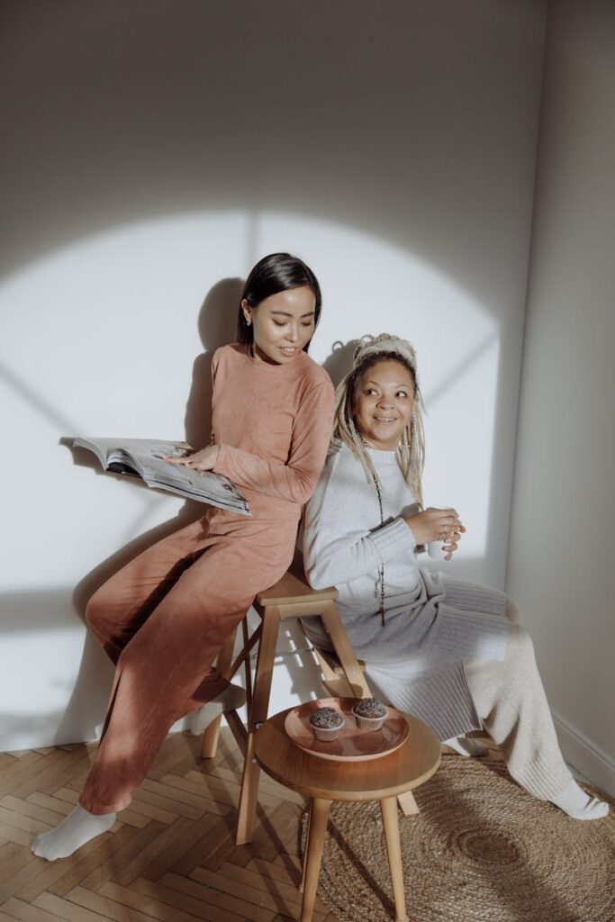 Two women in loungewear enjoy morning sunlight indoors, sitting and reading.