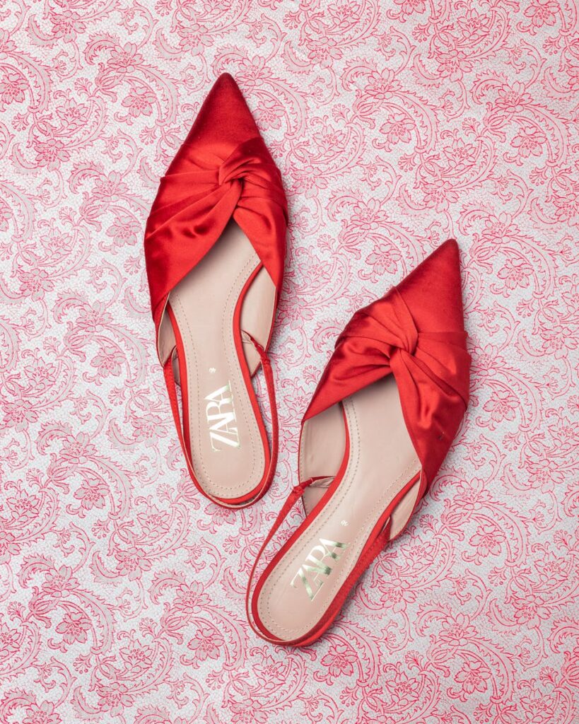 Close-up of red satin mules with a twisted design on an ornate patterned background for fashion photography.