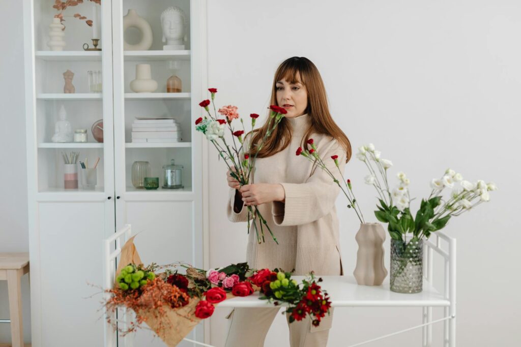A woman arranging flowers in vases in a bright indoor space, creating a floral arrangement.