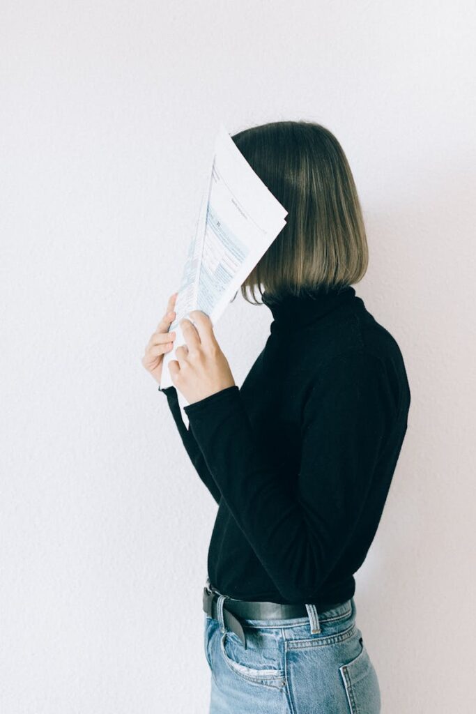 A woman indoors holding paper to conceal her face against a white wall background.