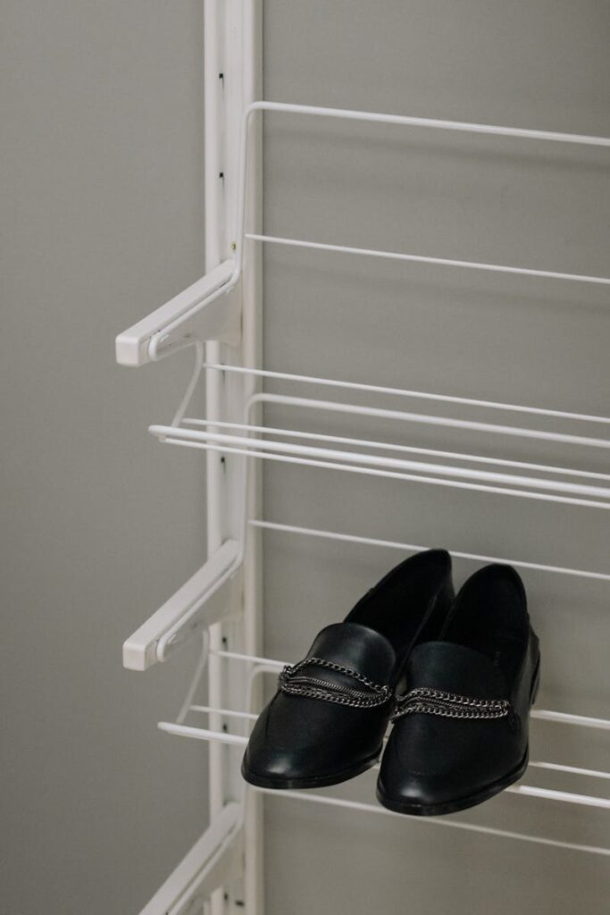 Pair of black leather shoes with chain detail on a minimalist white shelf, indoors.