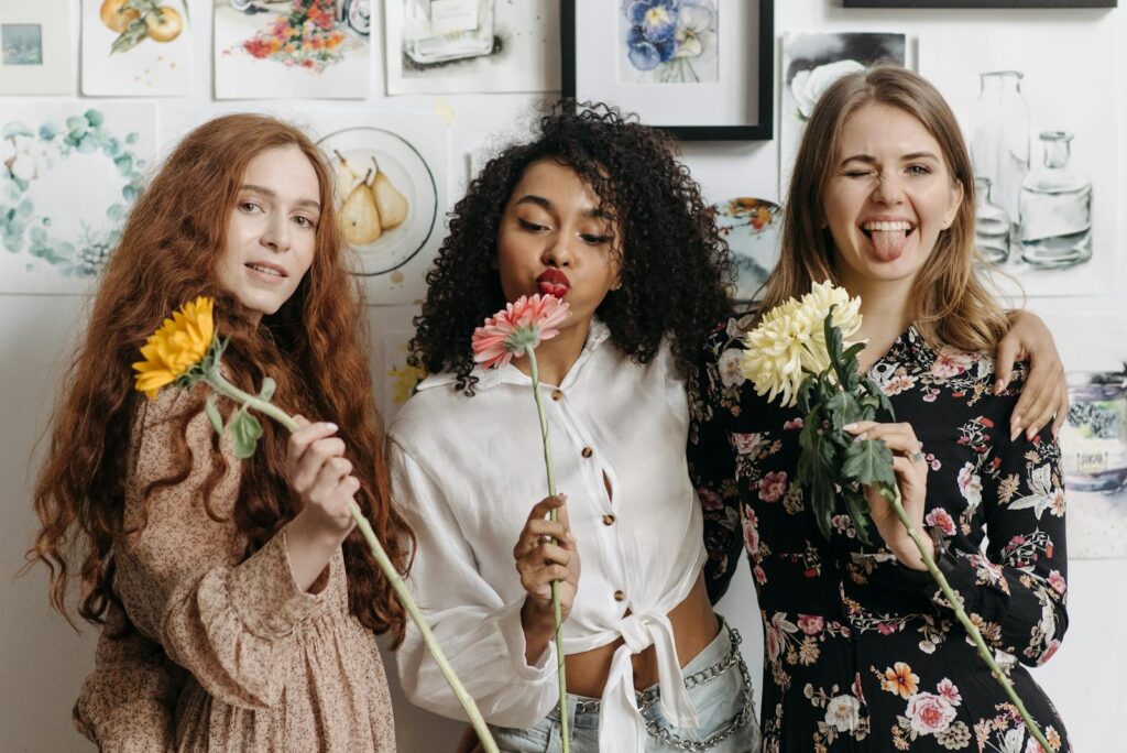 Three friends enjoy a fun moment posing with flowers against an artistic wall.